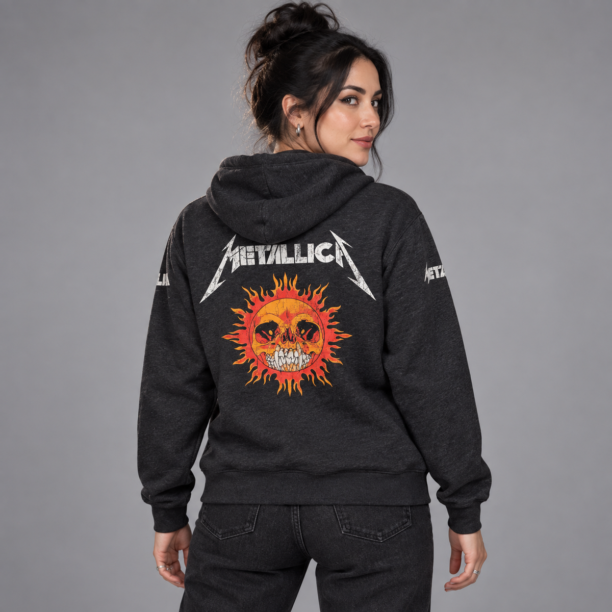 Studio back view of a woman wearing the charcoal Metallica Worn Pushead hoodie, showing the orange sun-skull and white band logo.